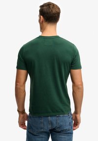 Un homme portant un t-shirt vert foncé à manches courtes et un jean bleu, debout de dos face à l'appareil photo sur un fond blanc uni.