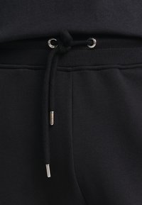 Primo piano della vita di pantaloni sportivi neri con cordino nero legato infilato attraverso occhielli metallici e terminali metallici alle estremità.