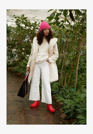 Kreemivärvi puffer vest, beežist kudumist kampsunist, valgest laia säärtega pükstest, erksatest punastest kingadest ja roosast beanie-mütsist. Must käekott punase ja valge rihmaga.