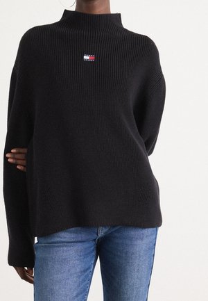 Kobieta nosząca czarny prążkowany sweter Tommy Jeans z małą naszywką z logo na piersi, zestawiony z niebieskimi dżinsami.