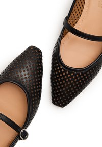 Bailarinas Mary Jane de cuero negro perforado con punteras puntiagudas y correas en el tobillo, plantillas beige y una hebilla plateada en el zapato izquierdo.