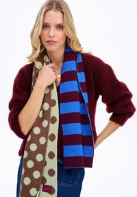 Pletené šály: jeden hnedý so zelenými bodkami, druhý vínový s modrými pruhmi; nosené cez vínový chlpatý sveter.