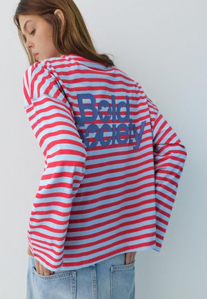 Mladá osoba v modro-červenej pruhovanej dlhorukávovej košeli s textom „Bold Society“ na chrbte, doplnená svetlými denimovými džínsami.