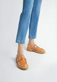 Mocassini in pelle color tan con una fibbia dorata e una robusta suola beige. Abbinati a jeans in denim blu con risvolto.