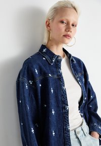 Camisa de ganga em azul escuro com bordado floral branco, frente com botões e colarinho clássico. Usada sobre uma blusa branca de canelado.