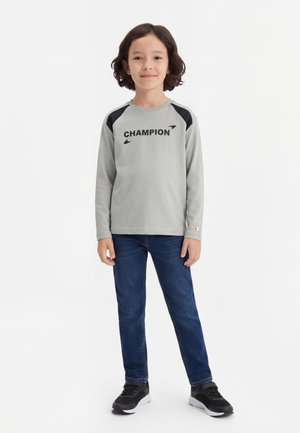 Dieťa s tmavými vlasmi po plecia stojí a usmieva sa, má na sebe sivé tričko s dlhým rukávom "Champion", modré džínsy a čierne tenisky.