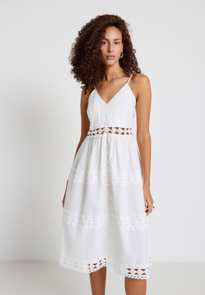 Donna con capelli ricci che indossa un abito midi bianco senza maniche con inserti in pizzo, dettagli con bottoni e scollo a V, in piedi davanti a uno sfondo semplice.