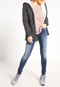 Grå huva jacka med vit fleecefoder, ljusrosa t-shirt, blå denimjeans och grå sneakers. Slätt tyg, avslappnad design.