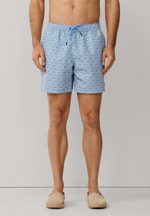 Hombre con bañador azul estampado con cordón y alpargatas beige sin cordones, de pie contra un fondo liso.