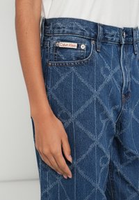 Jeans de ganga azul com padrão acolchoado em forma de diamante, com um emblema vermelho do logo Calvin Klein, ferragens metálicas e design padrão com cinco bolsos.