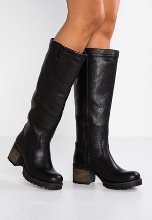 Personne portant des bottes noires en cuir jusqu'au genou avec des talons épais et des semelles texturées, debout sur un sol blanc devant un fond clair.