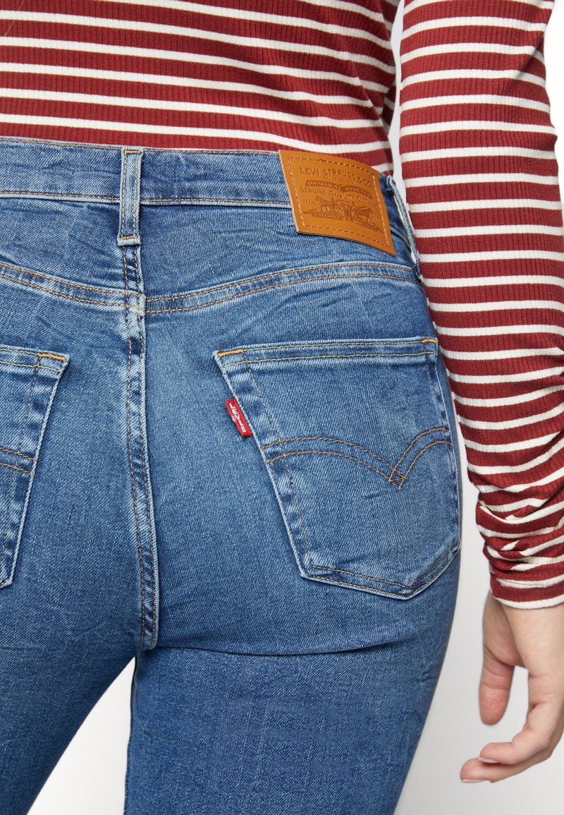 Blaue Denim-Jeans mit hoher Taille, ausgestattet mit orangefarbener Naht, zwei Gesäßtaschen und einem braunen Lederpatch am Bund.