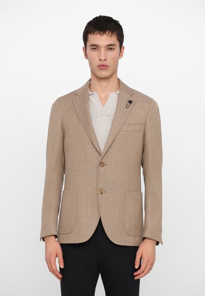 Light tan blazer with textured fabric, two buttons, and patch pockets; black accessory pinned on lapel; worn over a gray shirt.