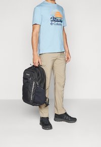 Hombre con camiseta gráfica azul, pantalones beige y zapatos de senderismo negros, sosteniendo una mochila negra de Columbia por su asa.