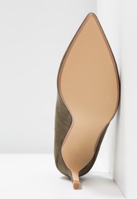 Zelená semišová lodička na vysokém podpatku s špičatou špicí a štíhlým podpatkem. Hladká nahá podrážka s texturovanými gripovými proužky. Detail bočního švu.