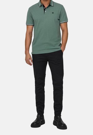 Grünes Poloshirt mit schwarzem Kragen, weißen Streifen und einem kleinen Logo. Kombiniert mit schwarzen Hosen und schwarzen Schuhen.