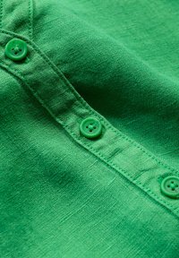Tessuto verde con una trama texturizzata, caratterizzato da una fila di bottoni verdi abbinati lungo il bordo. La vista ravvicinata mette in evidenza i dettagli del materiale.