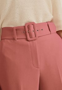 Pantalons ajustés roses avec une large taille, munis d'une boucle rectangulaire et de passants de ceinture. Tissu lisse avec une texture subtile et poches avant.