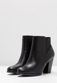 Et par sorte læder ankelstøvler med blokhæle og elastiske sidestykker på en hvid overflade mod en ensfarvet lys baggrund.