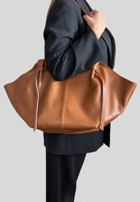 Velká hnědá kožená taška typu tote s texturovaným povrchem, staženými boky a širokým otvorem. Má pevné ramenní popruhy.