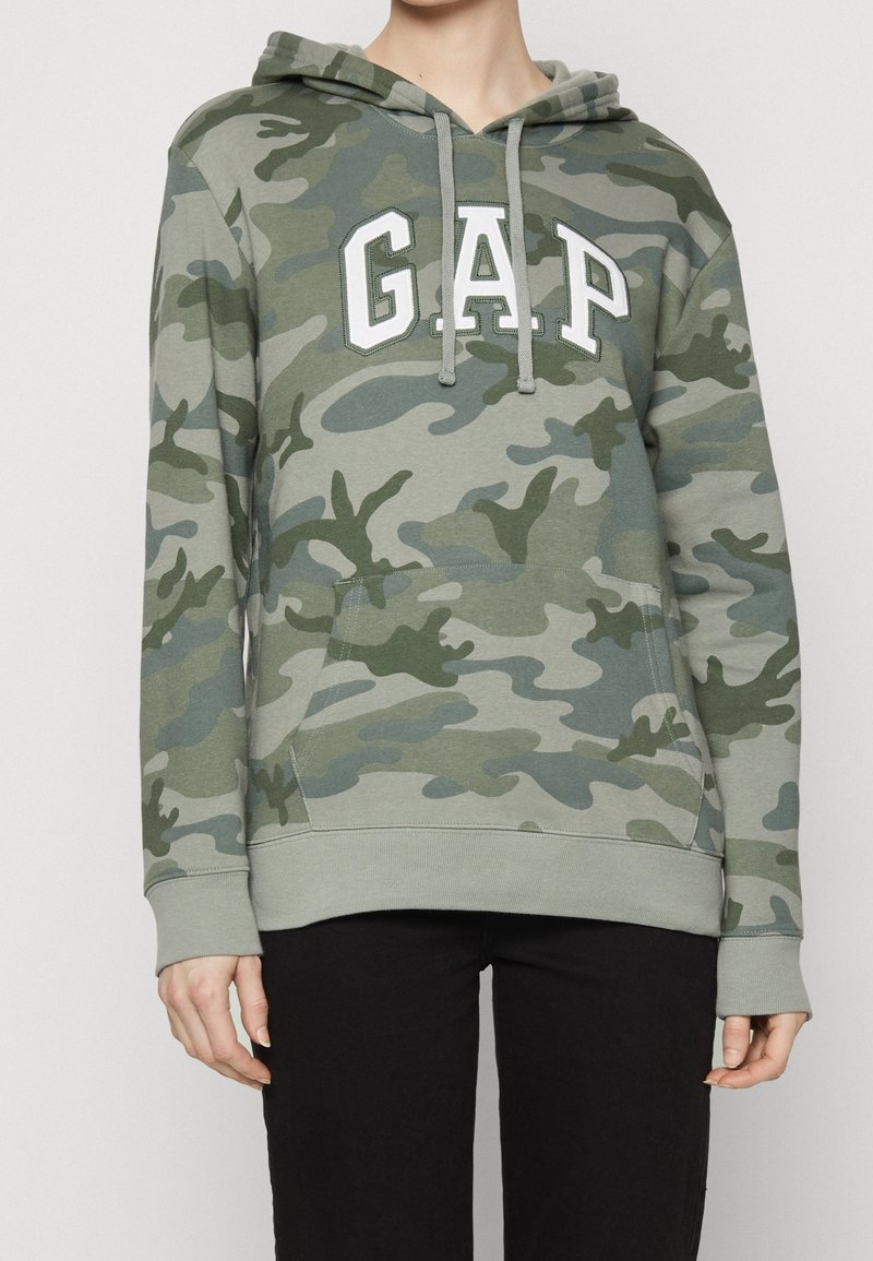 Persona con sudadera con capucha de camuflaje verde y gris con el logo "GAP" en blanco en el pecho y pantalones negros, de pie contra un fondo liso.