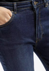 Granatowe dżinsy o gładkiej teksturze, z kontrastowym pomarańczowym przeszyciem, metalowym zapięciem na guzik oraz pięcioma kieszeniami.