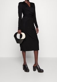 Schwarzes Wickelkleid mit langen Ärmeln, V-Ausschnitt und taillierter Passform. Kombiniert mit schwarzen High-Heel-Mules und einer schwarz-weißen runden Handtasche.