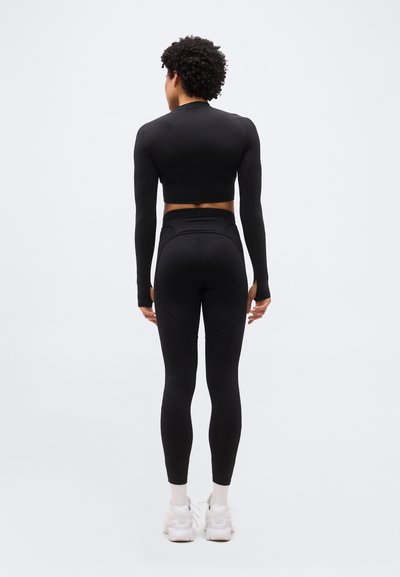 Άτομο με σγουρά μαλλιά στέκεται προς τα πίσω φορόντας μαύρο crop top με μακριά μανίκια, κολάν με ψηλή μέση, λευκές κάλτσες και λευκά αθλητικά παπούτσια σε απλό φόντο.