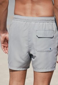 Shorts de bain gris en tissu léger avec une taille élastique et une poche arrière dotée d'un bouton. Texture lisse, coupe au-dessus du genou.