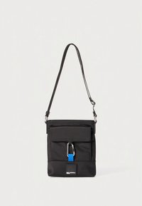 Mala preta de tecido para usar ao peito, com fecho de correr no topo, bolso frontal com aba, ferragens prateadas e detalhe azul. Apresenta uma alça ajustável.