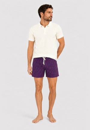 Mann in weißem kurzärmligem Poloshirt und blauen, gemusterten Shorts mit Kordelzug steht barfuß vor einfarbig hellem Hintergrund.