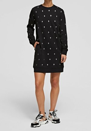 Personne portant une longue robe-sweat noire avec de petits motifs de crânes blancs, mains dans les poches, associée à des baskets épaisses blanches et noires.