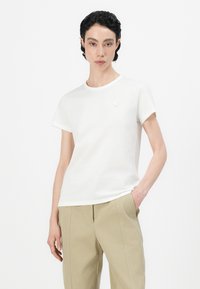 Λευκό βαμβακερό μπλουζάκι με κοντά μανίκια, στρογγυλή λαιμόκοψη και μια μικρή κεντημένη λεπτομέρεια στο στήθος, συνδυασμένο με μπεζ παντελόνι.