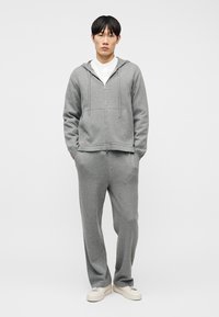 Νέος άνδρας όρθιος με τα χέρια στις τσέπες, φορώντας γκρι φούτερ με φερμουάρ, παντελόνι φαρδύ σετ, λευκό πουκάμισο και λευκά αθλητικά παπούτσια σε απλό φόντο.