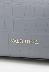 Superficie de cuero texturizado en gris con un patrón de cocodrilo en relieve y letras doradas metálicas que dicen "VALENTINO" centradas en la tela.