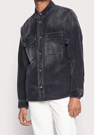 Hombre vestido con una camisa de botones de pana gris oscuro con dos bolsillos en el pecho y pantalones blancos frente a un fondo simple.