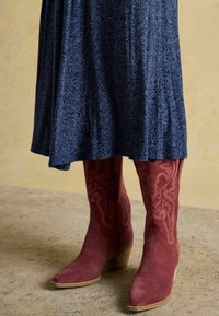 Sinine mustriga seelik langeb punaste sametist kauboi saapadele, millel on detailne õmblus, ning on asetatud tekstuursele betoonpinnale.