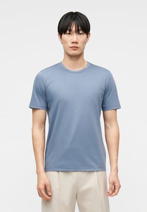 Uomo in piedi, volto frontale, che indossa una maglietta blu a maniche corte semplice e pantaloni beige, con uno sfondo chiaro uniforme.