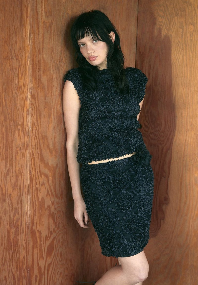 Mustakirjuline kaheosaline komplekt, kus on tekstureeritud lühikeste varrukatega ülahern ja sobituv seelik, millel on dekoratiivne aksessuaar.