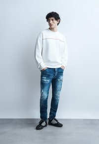 Jonge man staand met handen in de zakken, gekleed in een witte trui, versleten blauwe spijkerbroek en zwarte sneakers met witte strepen, tegen een effen witte muur.