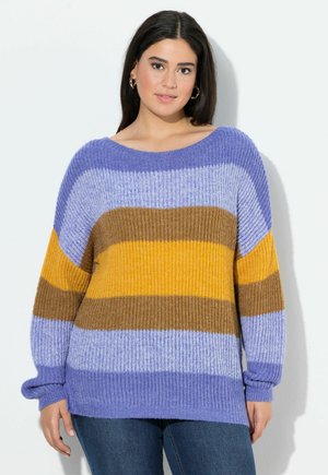 Junge Frau mit goldenen Creolen und einem lockeren Strickpullover in lila, braun und gelben horizontalen Streifen, die vor einer weißen Wand steht.