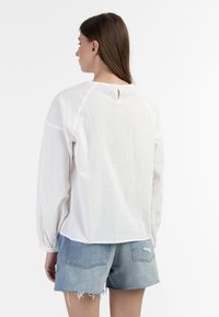 Weiße Baumwollbluse mit langen Ärmeln, lässigem Schnitt und einem kleinen Schlüsselverschluss auf der Rückseite. Kombiniert mit hellblauen Denim-Shorts.
