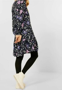 Blommig svart klänning med långa ärmar och volangkant, prydd med lila och blå blommönster, kombinerad med svarta leggings och vita högtoppsneakers.