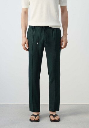 Man draagt donker groene rechte broek met koord in de taille, wit gestructureerd kortarmig hemd en zwarte teenslippers, staand binnen.