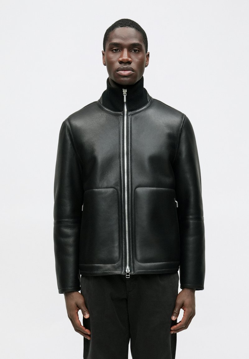 Schwarze Lederjacke mit hohem Kragen, Frontreißverschluss und zwei großen Vorder Taschen. Glatte Textur und minimalistisches Design.