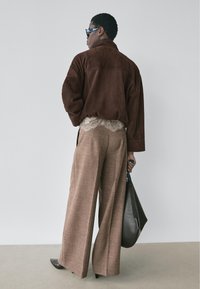 Persoon draagt een oversized bruine suède jas, top met kantafwerking, wijde bruine broek, zwarte hakken en houdt een zwarte handtas vast, met de rug naar de camera gericht.
