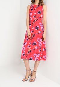 Abito senza maniche rosa con stampa floreale vivace, lunghezza al ginocchio, tessuto liscio. Abbinato a sandali con tacco a strati argento e nero con motivo.