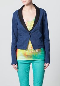 Mörkblå, kort blazer med svart sjalkrage, knappstängning med en enkel knapp, och långa ärmar över en flerfärgad tie-dye tanktopp och gröna byxor.