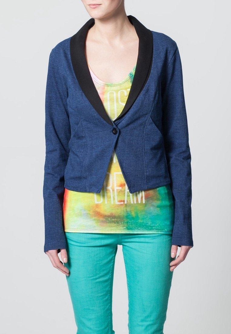 Mörkblå, kort blazer med svart sjalkrage, knappstängning med en enkel knapp, och långa ärmar över en flerfärgad tie-dye tanktopp och gröna byxor.