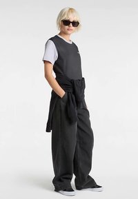 Haut noir sans manches porté sur un t-shirt gris, pantalon large noir et baskets noires. Accessoires : lunettes de soleil foncées et pull noir noué.
